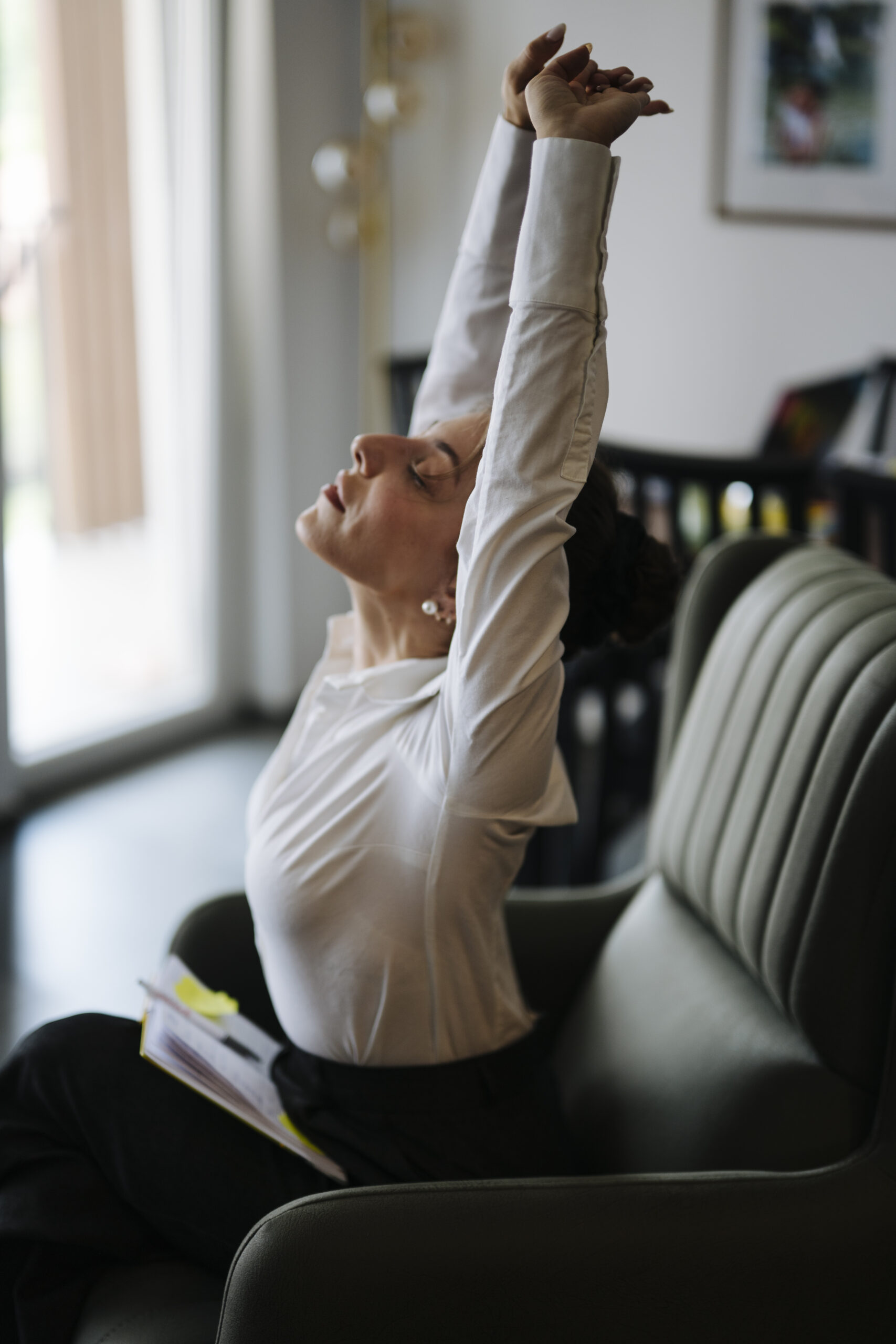 Woman seated in an upholstered chair with her hands clasped and arms reaching overhead.